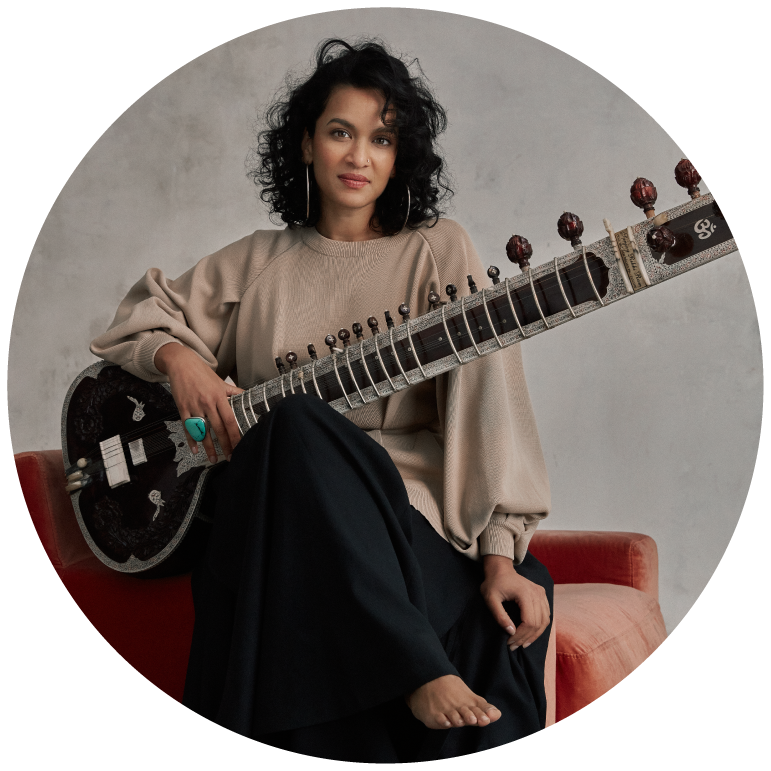Anoushka Shankar wears a beige top and black pants and poses with her sitar against a grey backdrop
