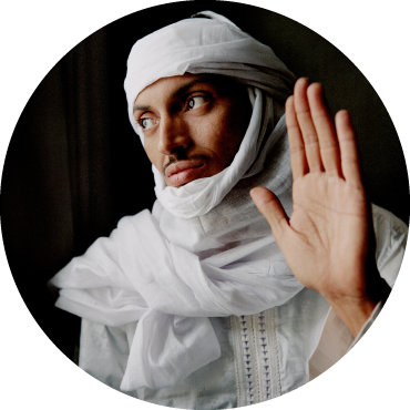 Bombino wears white clothing and looks to the side with his hand held up against a black backdrop