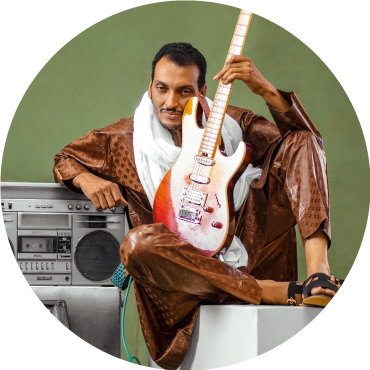 Bombino wears brown clothing and poses with his guitar against a boombox in front of a green backdrop