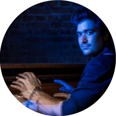 In dim blue lighting, Harold López-Nussa plays the piano and swivels his head towards the camera