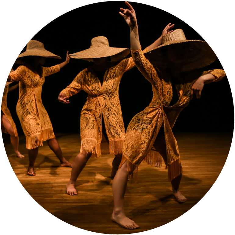 Three dancers are on stage. They have dark skin and wear orange, lacy robes, and straw hats.