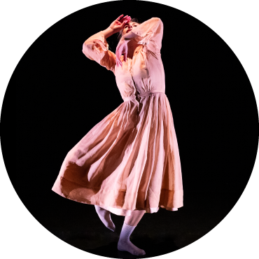 Dancer wears a light pink dress and is captured in motion against a black backdrop