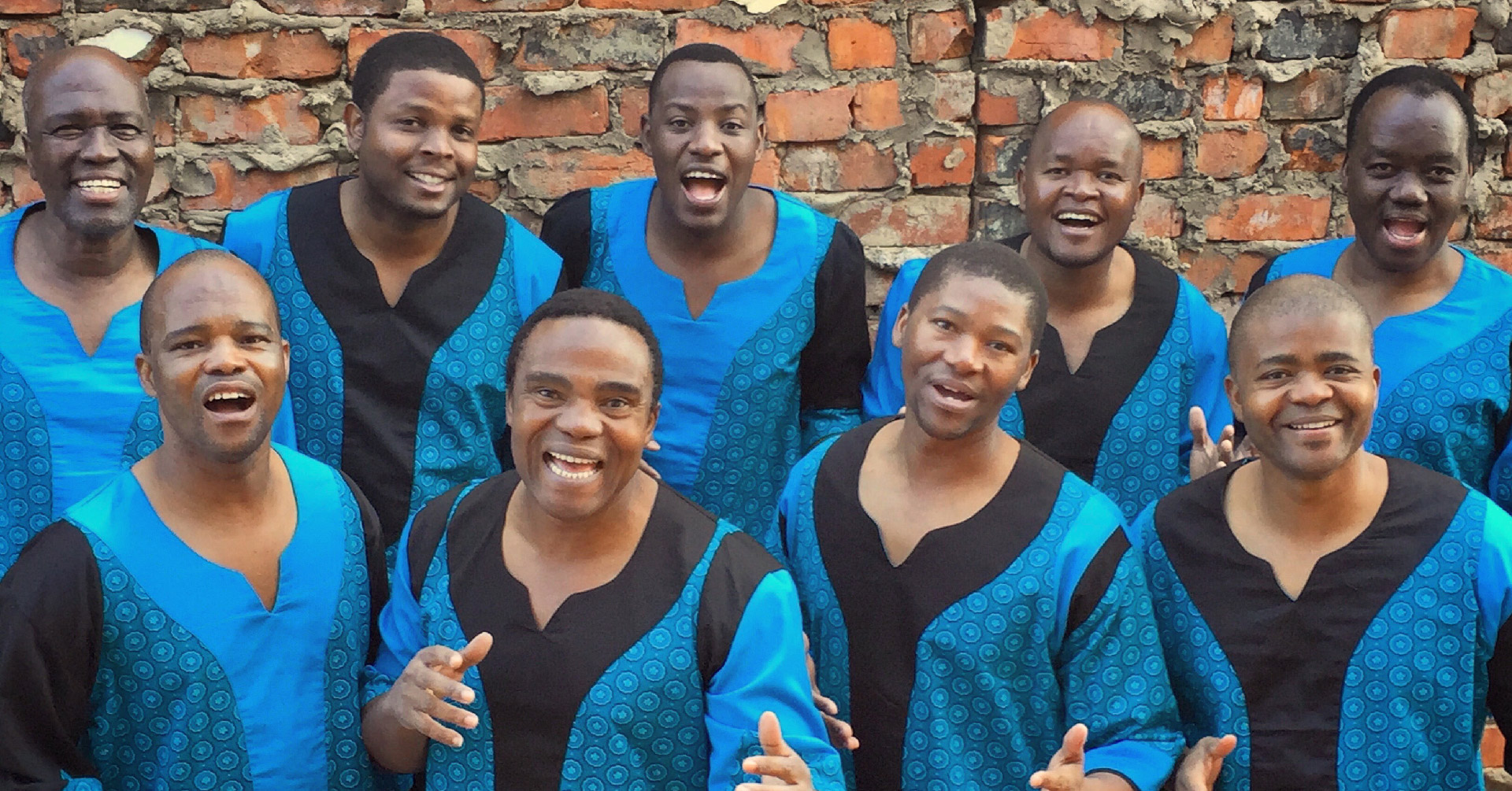 Members of Ladysmith Black Mambazo look at the camera and stand in front of a red brick wall. They wear blue and black robes. 