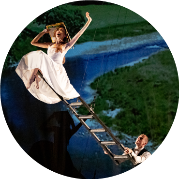 A woman sits at the top of a ladder with her arms raised above her wearing white as a man stands at the bottom of the ladder in front of a blue and green screen