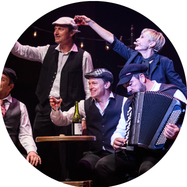 Members of the cast are dressed in black and white and hold glasses of wine as they perform live on stage