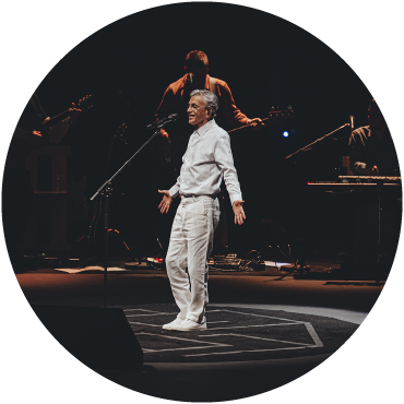 Caetano Veloso wears a white shirt and pants and is captured performing live on stage