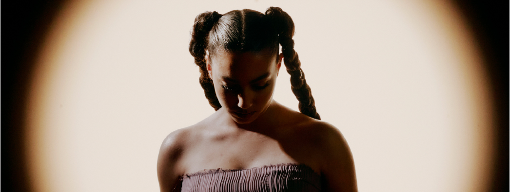 The singer stands waring a strapless pink dress, hair in braided pig tails, looking down against an off white backdrop