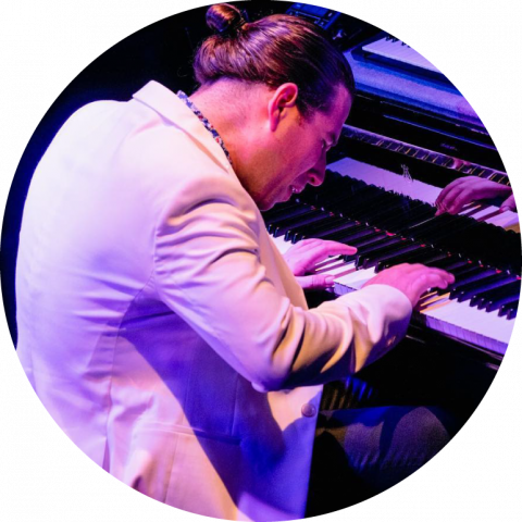 Alfredo Rodríguez wears a white blazer and plays piano live in blue lighting 