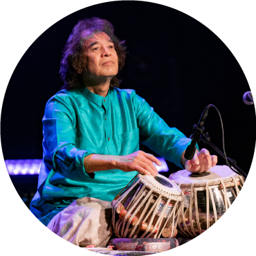 Zakir Hussain wears blue clothing and plays Table live on stage against a black backdrop
